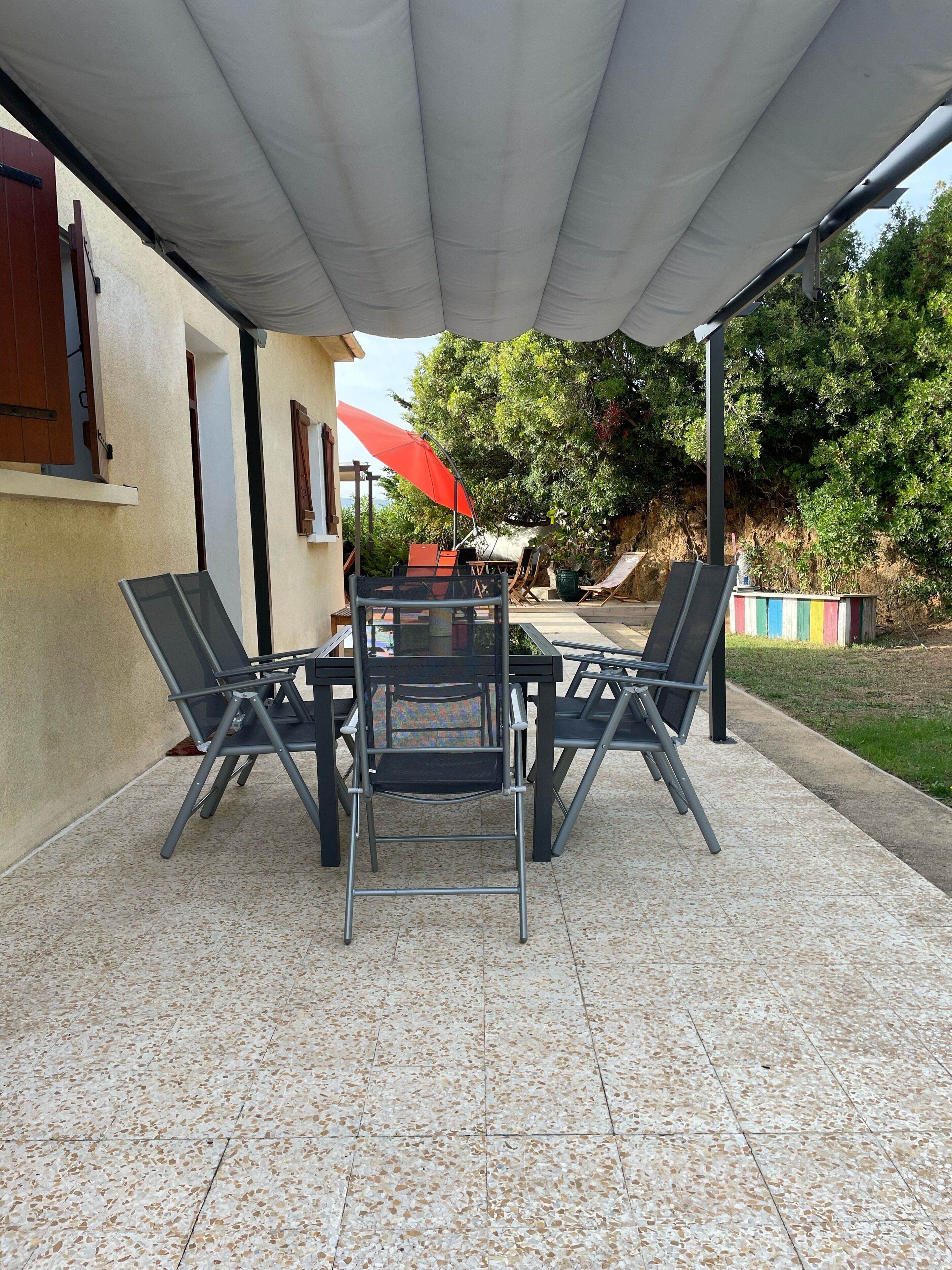 Un patio al aire libre con una mesa y sillas, un paraguas rojo y vistas a árboles y un edificio.