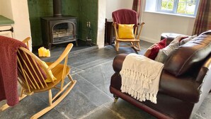 A living room with a stone fireplace, a rocking chair, a leather sofa, and a window with curtains. - Charming Cottage for 5 Near Dartmoor, Beach, Pub (Holsworthy)