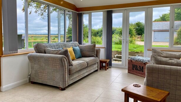 A spacious living room with large windows, a sofa, a small table, and a wicker basket. - Charming Cottage for 5 Near Dartmoor, Beach, Pub (Holsworthy)