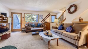A cozy living room with a sofa, coffee table, and TV. There is a staircase with a wreath on the wall. - Pinecreek #G- 3 BR: Close to Town- Shuttle to Slopes- Pool & Hot Tub Access (Breckenridge)