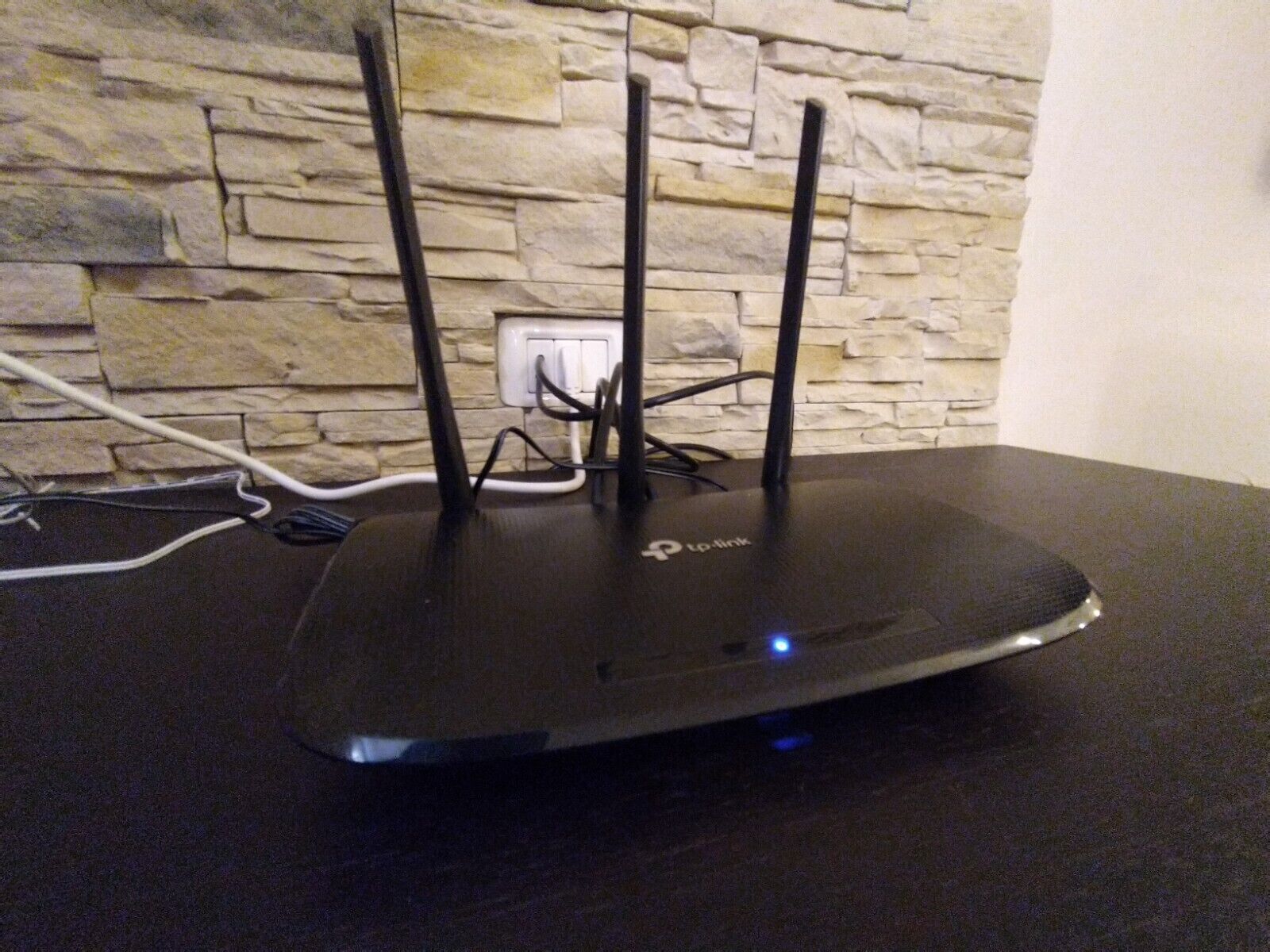 Ein schwarzer TP-Link Router mit drei Antennen auf einer schwarzen Oberfläche, verbunden mit einer weißen Steckdose.
