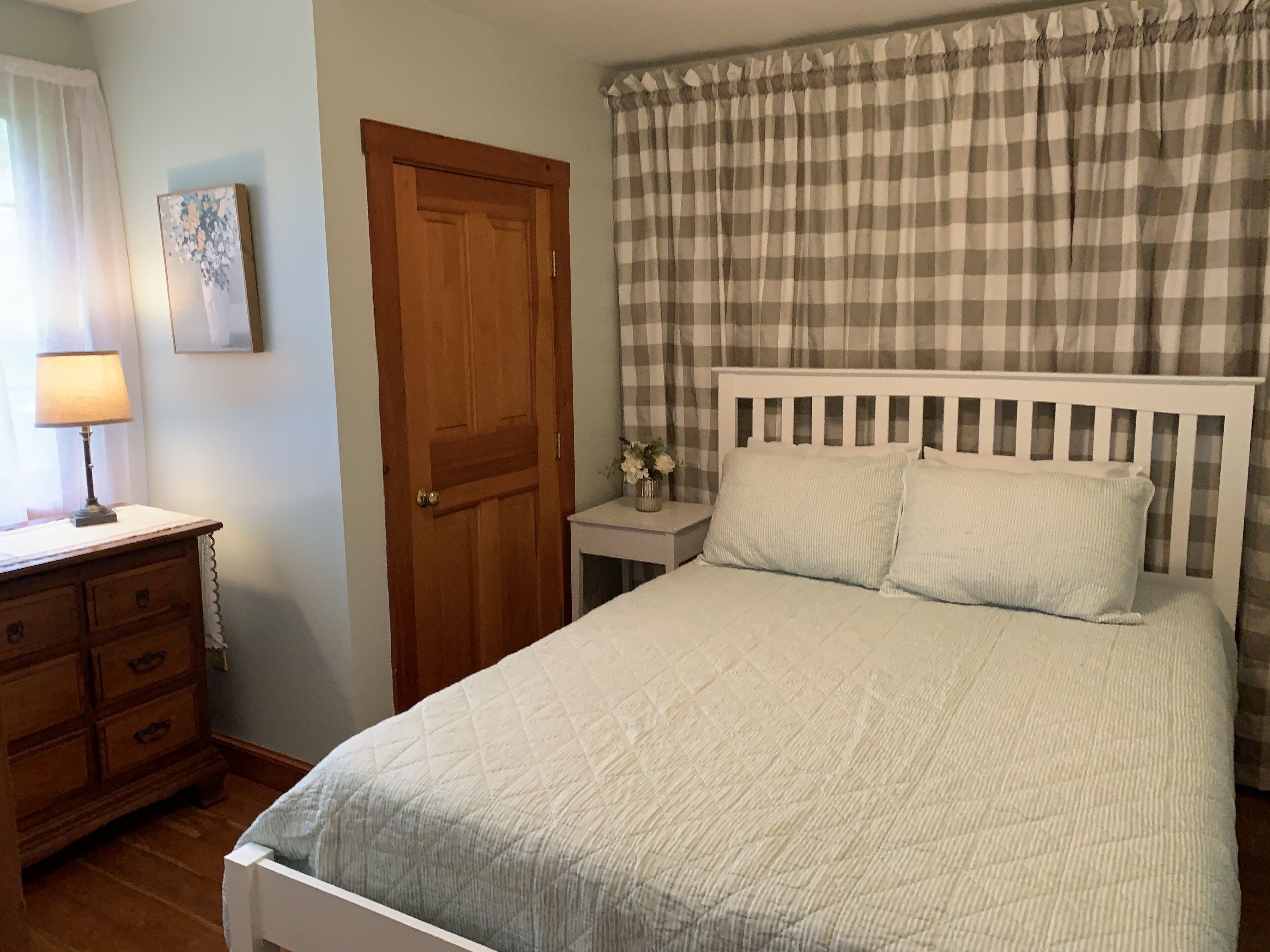 A bedroom with a bed, a wooden dresser, a nightstand with a lamp, a door, and a window with curtains.