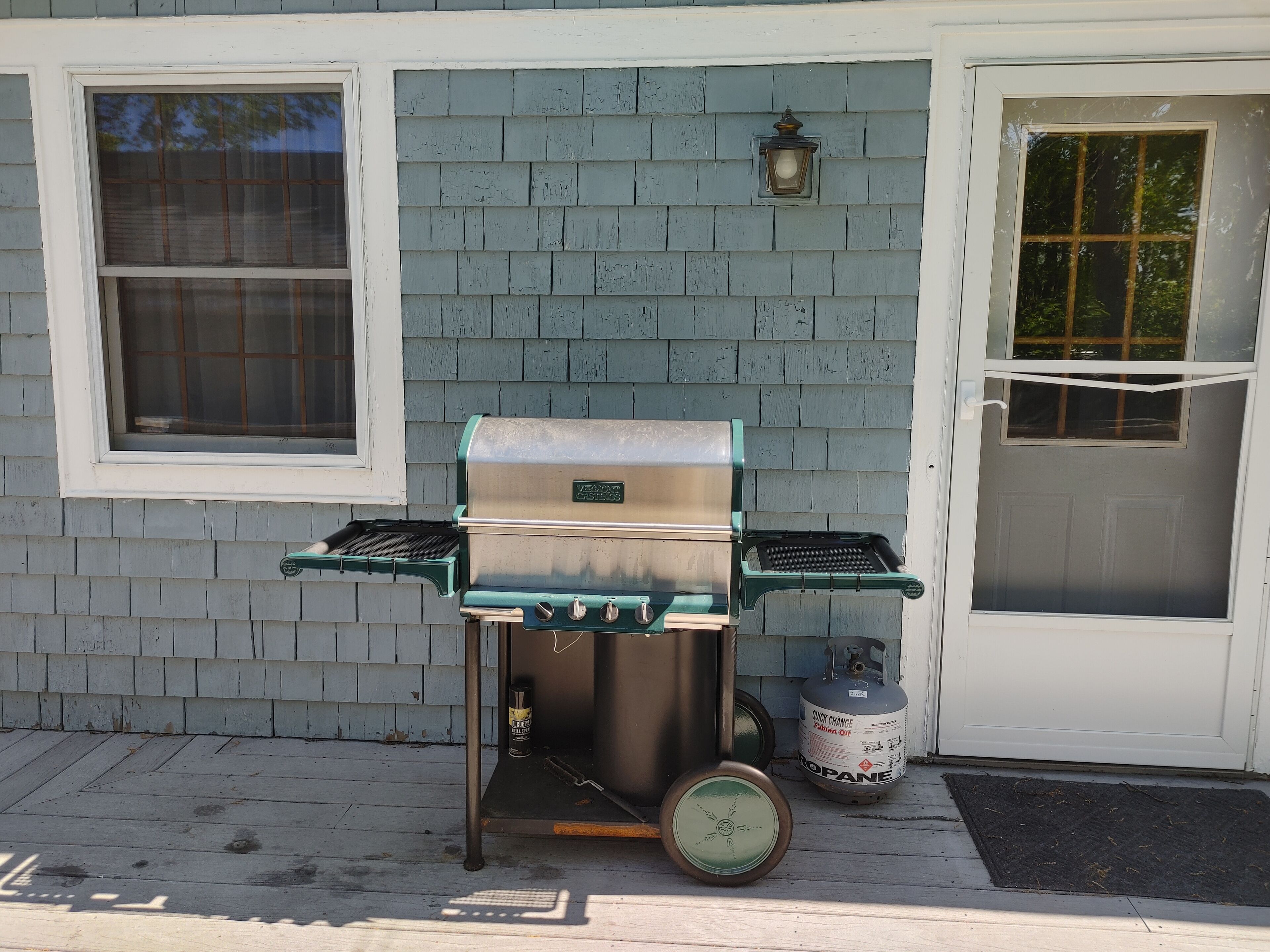 A portable grill with a propane tank, positioned on a porch with a white door and window.