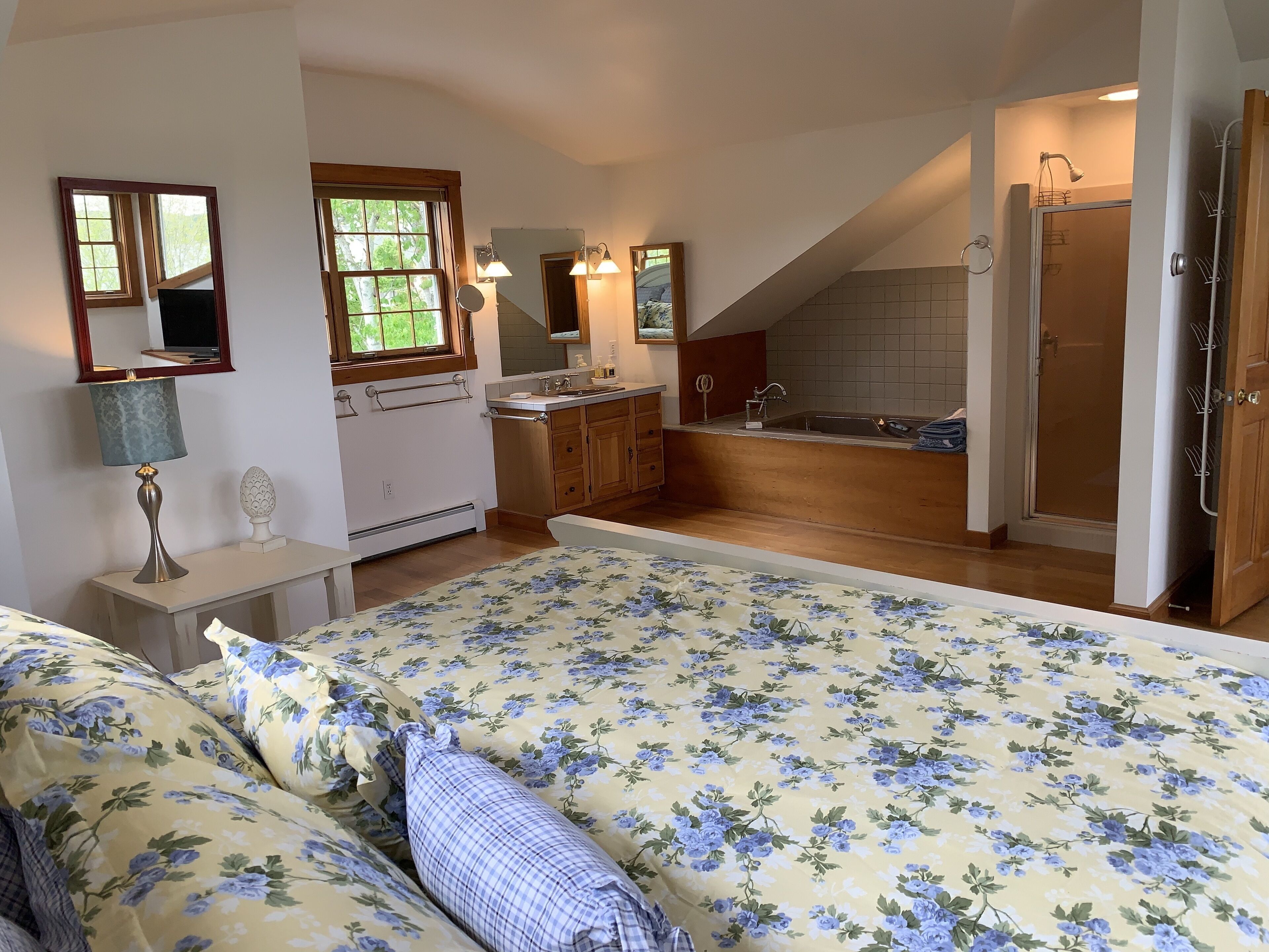 A bedroom with a large bed, a small bedside table, a lamp, and a window with a view of greenery.
