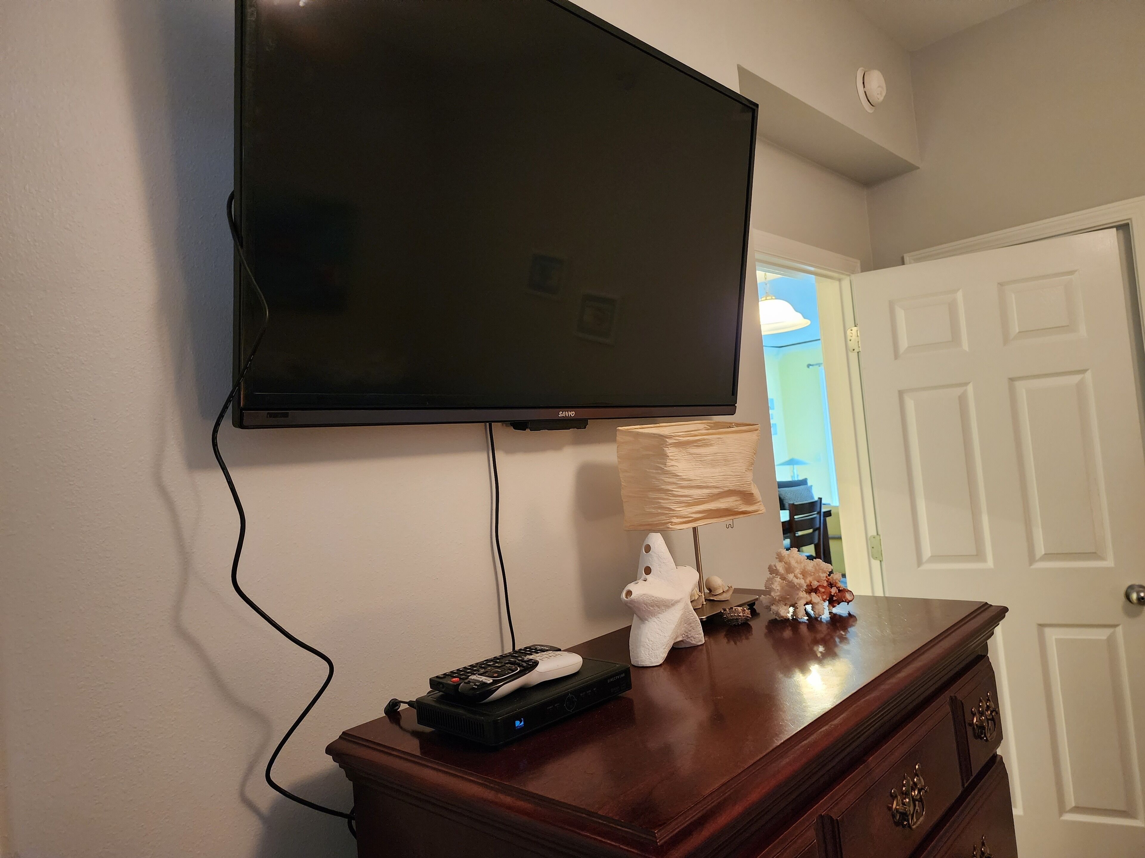 A television mounted on a wall, a wooden dresser with drawers, and a lamp on the dresser.