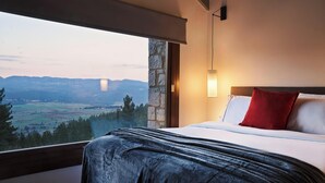 A bedroom with a large window, a bed with white linens and a red pillow, and a stone wall.