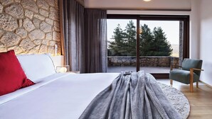 A bedroom with a stone wall, a bed with white and red pillows, a grey blanket, a green armchair, and a view of trees through a large window.