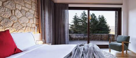 A bedroom with a stone wall, a bed with white and red pillows, a grey blanket, a green armchair, and a view of trees through a large window.
