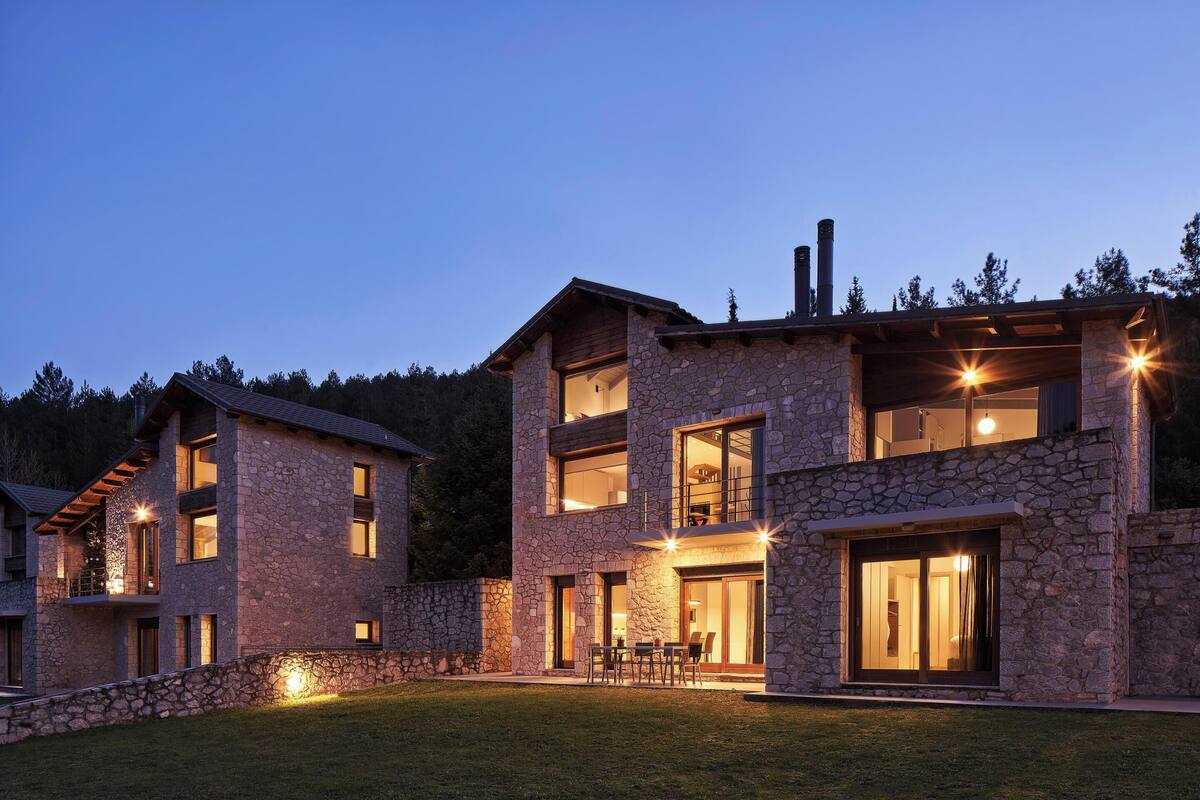 A modern two-story stone house with large windows and a well-lit outdoor area.