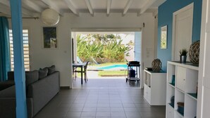 Ett rymligt vardagsrum med en soffa, ett matbord med stolar, ett köksområde med skåp och utsikt över en swimmingpool och grönområden utanför.