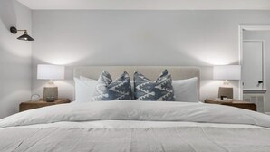Una cama bien hecha con ropa blanca, dos lámparas de noche y una almohada estampada.