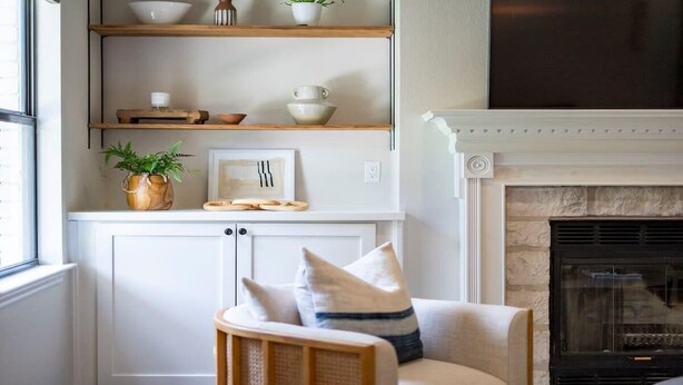 Un salón con chimenea, estanterías de madera con adornos, un sillón con cojines y una planta en maceta.