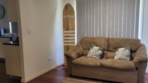 Ett vardagsrum med en beige soffa, två kuddar och en träinredd surfbräda som dekoration. I bakgrunden syns ett köksområde.