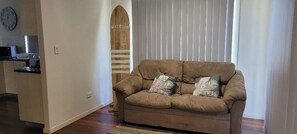 A living room with a beige sofa, two pillows, a wooden surfboard decoration, and a kitchen area in the background.
