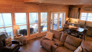 A cozy living room with a leather sofa, a dining table, and a kitchen area with wooden cabinets and large windows. - Beautiful cottage with AMAZING view of Barren River Lake! (Scottsville)