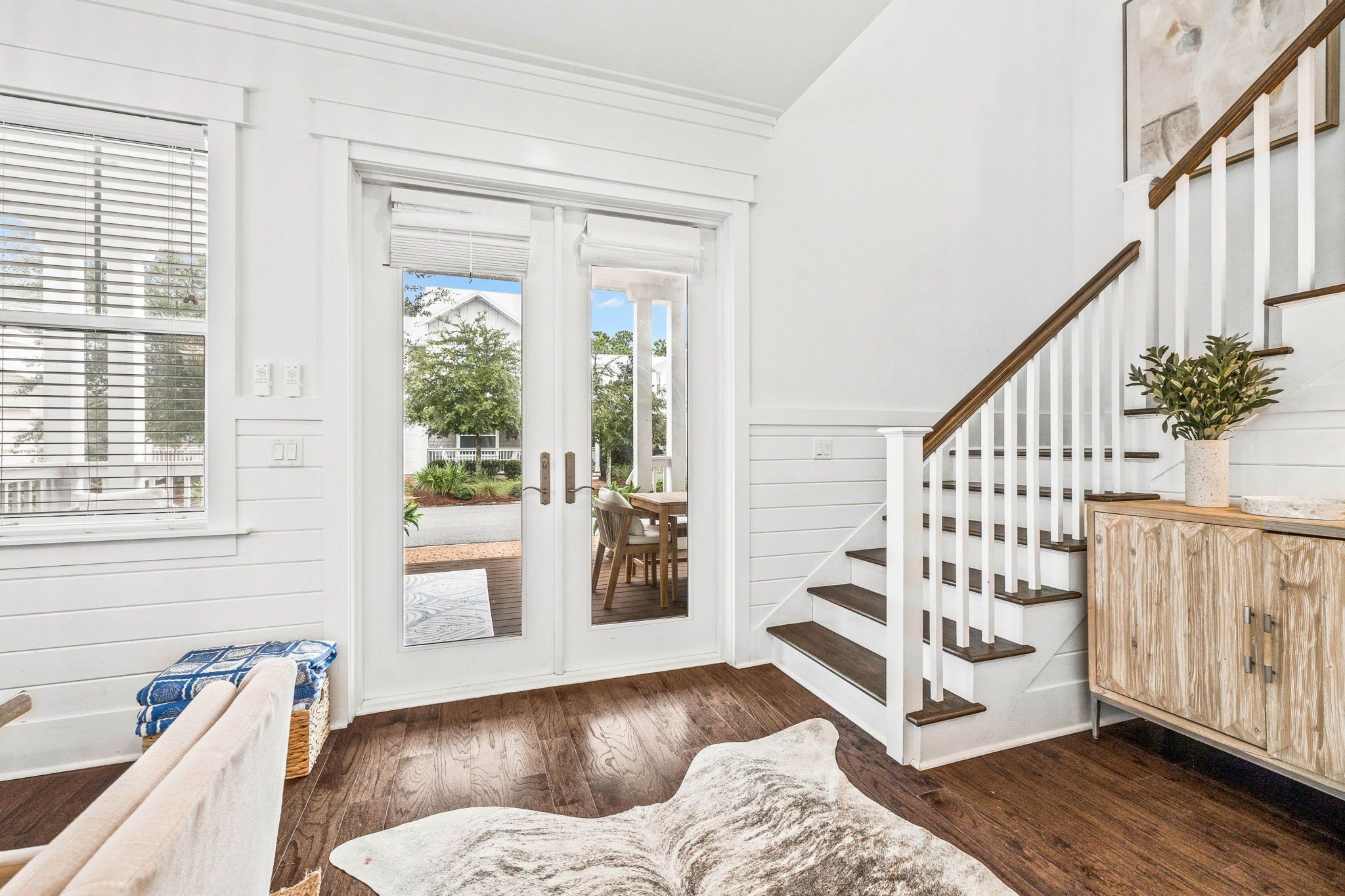 A spacious room with a wooden floor, a white door leading outside, a staircase with white railings, and a wooden cabinet with a plant on top.