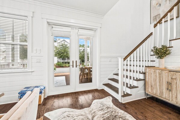 A spacious room with a wooden floor, a white door leading outside, a staircase with white railings, and a wooden cabinet with a plant on top.