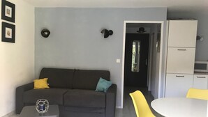 A living room with a dark grey sofa, a small side table, a yellow chair, and a white table. There is a kitchen area with a white refrigerator and cabinets.