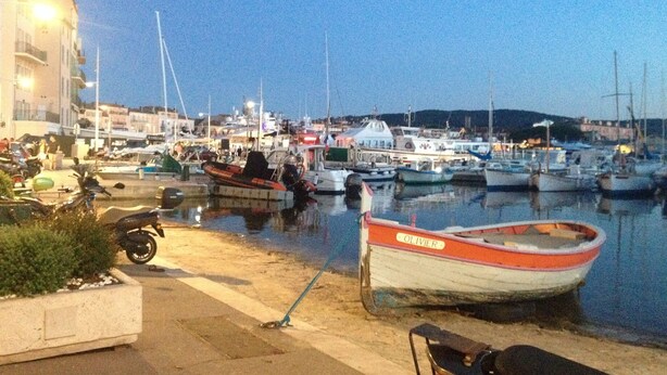 A marina with boats docked, a small boat named 'OLIVIER' in the foreground, and a building with a sign 'PARKING' on the left.