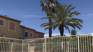 A swimming pool with clear blue water, surrounded by a white fence. In the background, there are palm trees and a building with a tiled roof.