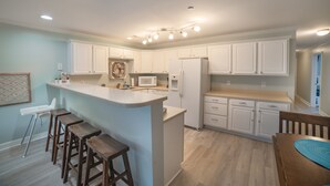 A modern kitchen with white cabinets, a central island with stools, a microwave, and a refrigerator. - BEACH PLEASE │ 2BR/2BA w/ 2 K in the heart of OKI! (Oak Island)