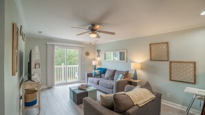 A living room with a sofa, a coffee table, a ceiling fan, and a large window. - BEACH PLEASE │ 2BR/2BA w/ 2 K in the heart of OKI! (Oak Island)