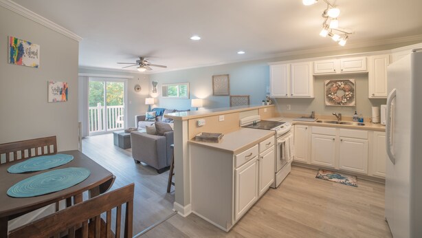 A modern kitchen with white cabinets, a central island, and a dining area with a wooden table and chairs. - BEACH PLEASE │ 2BR/2BA w/ 2 K in the heart of OKI! (Oak Island)
