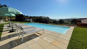 A swimming pool with lounge chairs and an umbrella, surrounded by a well-maintained lawn and a view of distant hills. - Tower House (Lapela)