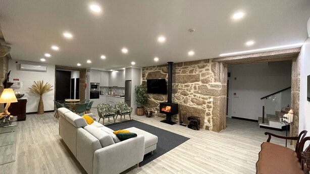 A modern living room with a stone fireplace, a sofa, a dining area, and a kitchen in the background. - Tower House (Lapela)