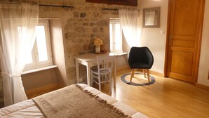 Um quarto com cama, escrivaninha, cadeira, porta de madeira e parede de pedra.