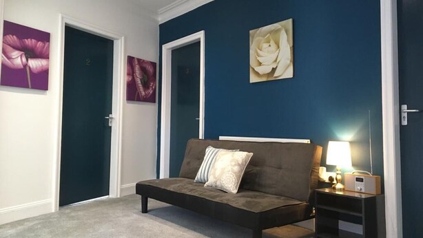 A room with a dark blue accent wall, a sofa, a bedside table with a lamp, and artwork on the walls. - En Suite Rooms at the Rose and Thistle (Berwick-upon-Tweed)