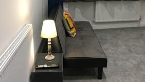 A modern bedroom with a grey sofa, a bedside table with a lamp, a remote control, and a pillow with a colorful design. - En Suite Rooms at the Rose and Thistle (Berwick-upon-Tweed)