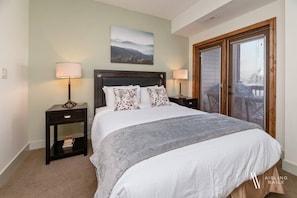 A bedroom with a bed, two bedside tables with lamps, a nightstand, a window with blinds, and a door with a glass panel. - The Sable Bright Side | Pool | Hot Tub | Top Floor (Radium Hot Springs)
