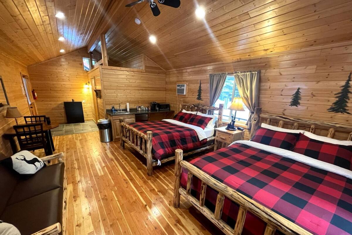 A cabin with wooden walls and floors, two beds with checkered bedding, a desk, a chair, and a small table.