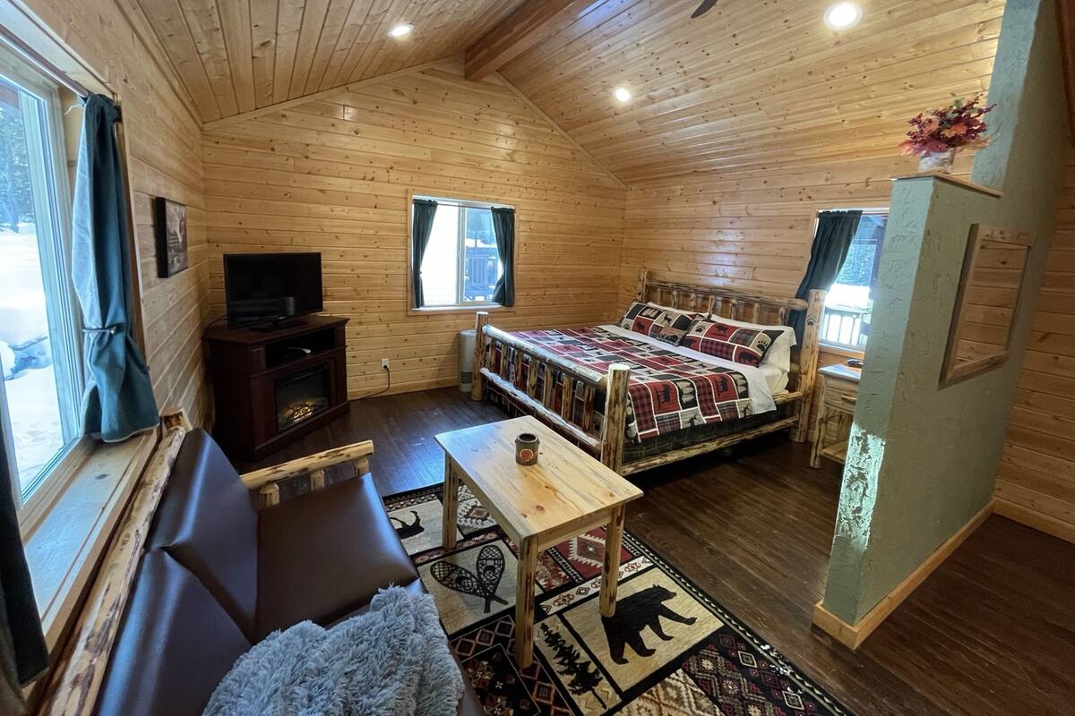 A cozy cabin interior with a wooden ceiling, a bed with a patterned blanket, a sofa, a small table, and a television.