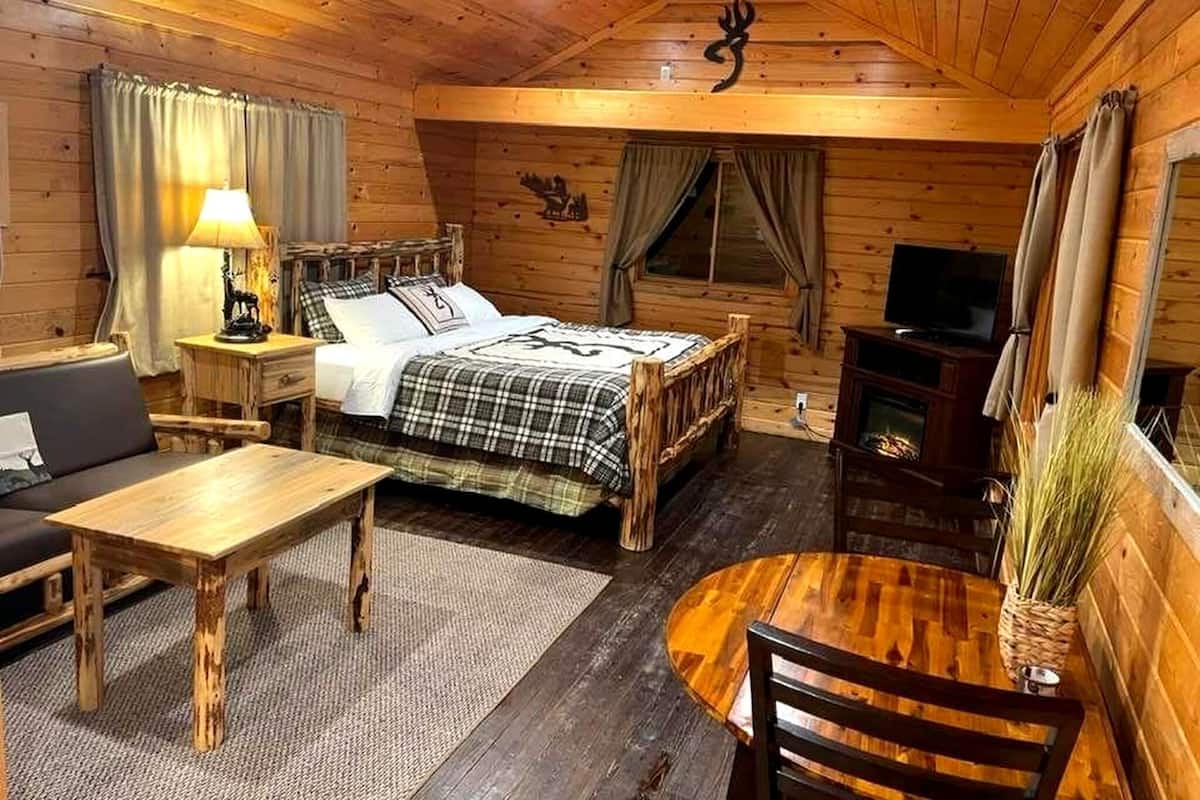 A wooden cabin interior with a bed, a sofa, a table, a TV, and a fireplace.