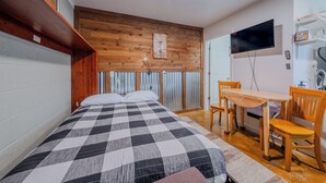 Camera da letto con copriletto a quadri, testiera in legno, pannello decorativo in legno a parete, televisore fissato al muro, tavolo in legno con sedie e una piccola mensola con oggetti.
