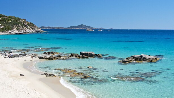 A beach with clear turquoise water, rocky outcrops, and a distant mountain. - Appartamenti Le Fontane con piscina (Villasimius)