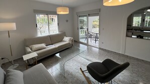 A modern living room with a sofa, a coffee table, a black chair, and a dining area with a table and chairs. There is a sliding glass door leading to a balcony with a view of greenery. - Vacation home in Oliva on the Costa Blanca, 10 minutes' walk from the sea. (Oliva Playa)