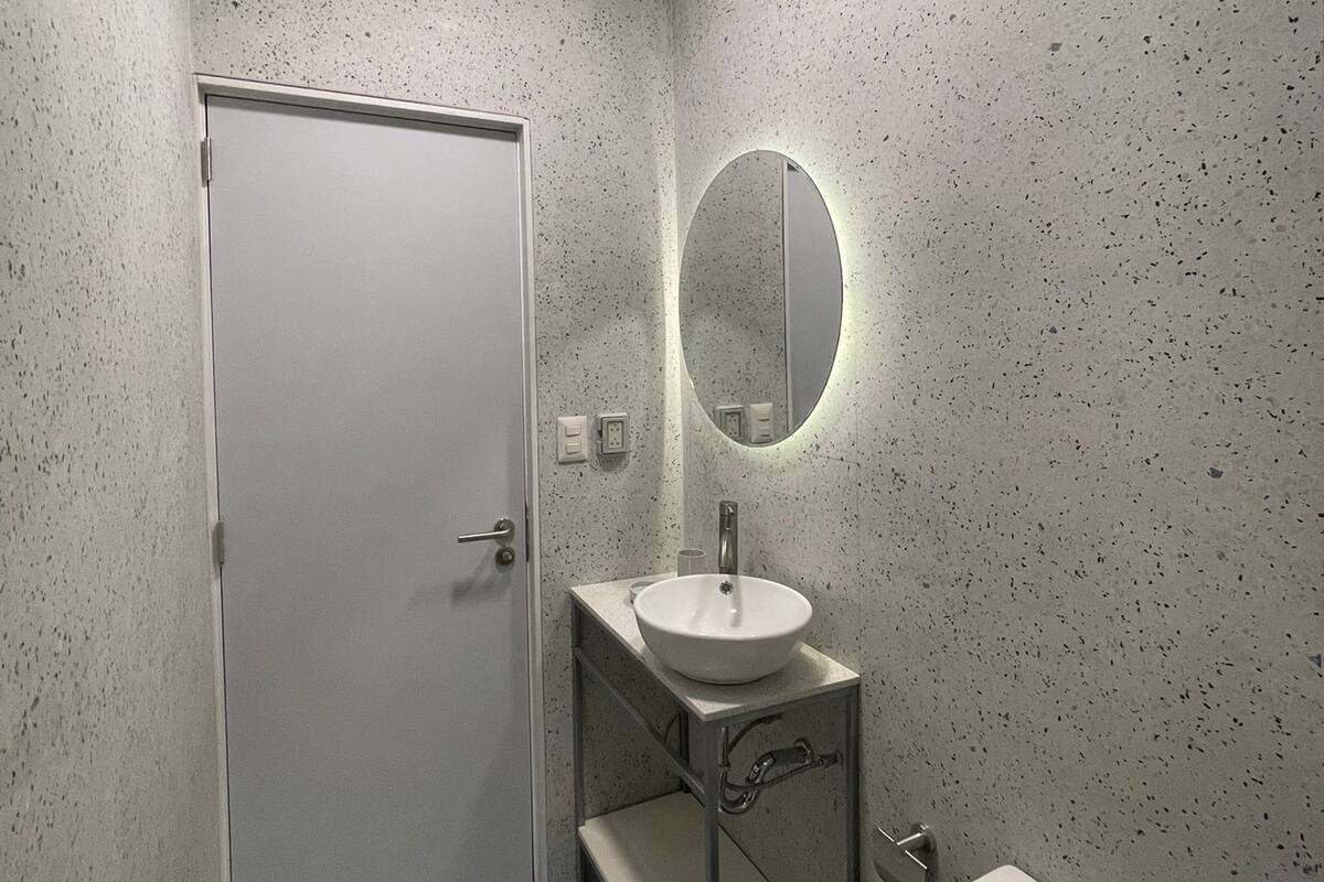 Un baño con una puerta blanca, un espejo redondo con luz, un lavamanos y un inodoro.