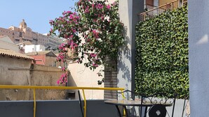 En balkon med et bord og stole, gelænder i gult, en væg med grøn vegetation, og en bygning med en kuppel i baggrunden.