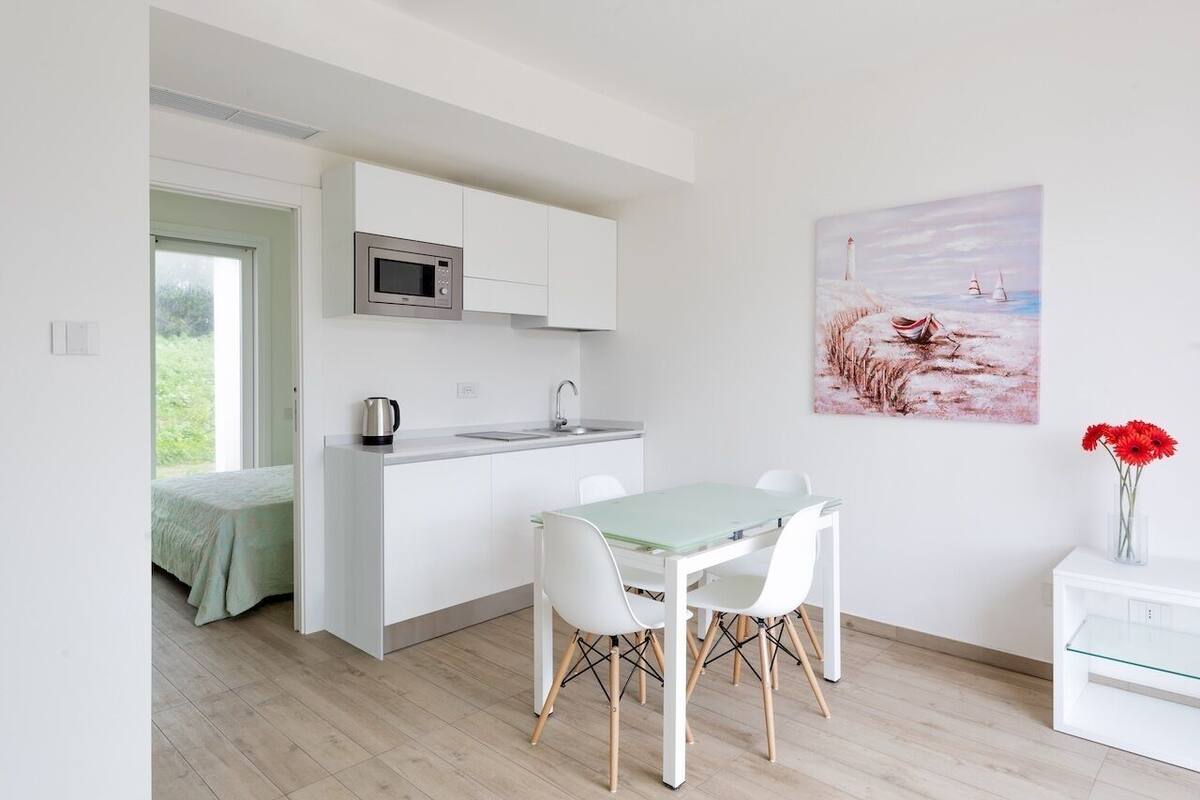 A modern kitchen with white cabinets, a microwave, and a sink. There is a dining area with a glass table and white chairs. A painting of a coastal scene is on the wall.