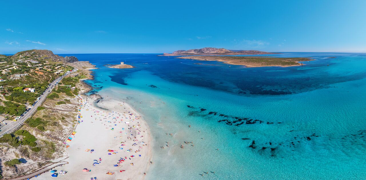 An aerial view of a coastal area with a beach, clear waters, and a road leading to the beach.