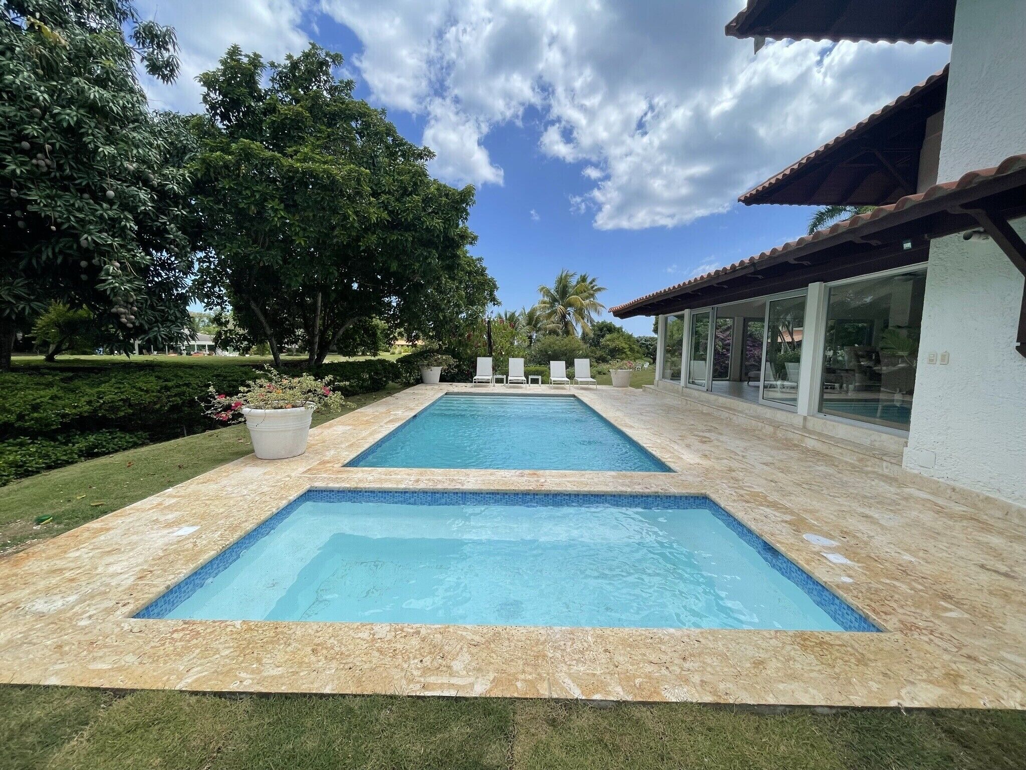 Una piscina rectangular con borde de azulejos, rodeada de césped cuidado y plantas en macetas.