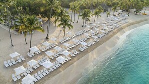 Une vue aérienne d’une plage avec des rangées de transats et de parasols blancs, des palmiers et un océan bleu limpide.