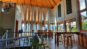 Un ambiente spazioso con soffitto in legno, ampie finestre con vista sulle montagne, zona pranzo con arredi in legno e angolo cottura.