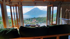 Un balcone con vista su una montagna e un lago. Si vede un letto con cuscini verdi e una poltrona sospesa.