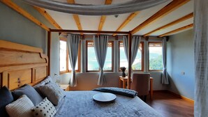 Camera da letto con testiera in legno, un letto con un vassoio rotondo in legno, un tavolino con un vaso e vista su un paesaggio montuoso attraverso ampie finestre con tende.