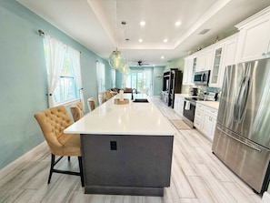 A modern kitchen with a central island, stainless steel appliances, and a dining area with a tufted chair. - Deluxe 3BR House with pool, short walk to beach (Cape Canaveral)