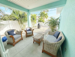 A covered patio area with wicker furniture, including chairs and a small table, surrounded by greenery.